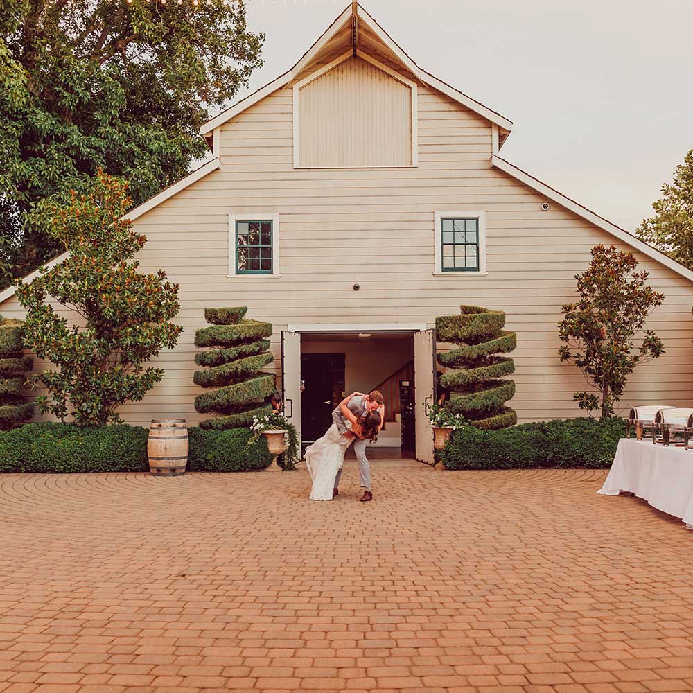 The groom dips his wife and kisses her as they stand in front of the 1918 barn.