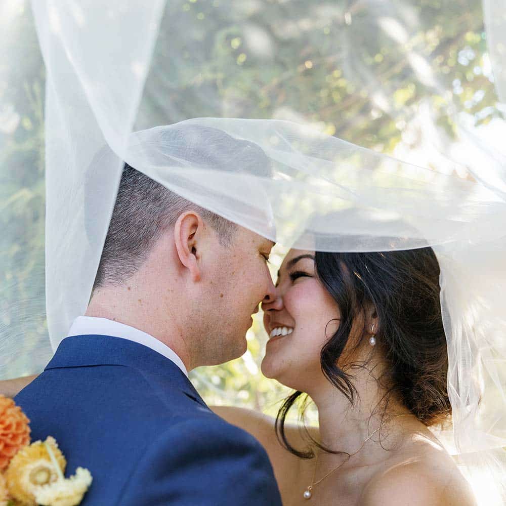 The bride and groom smile at each other as her veil partially covers their faces at their outdoor winery wedding in the Sacramento Wine Country.