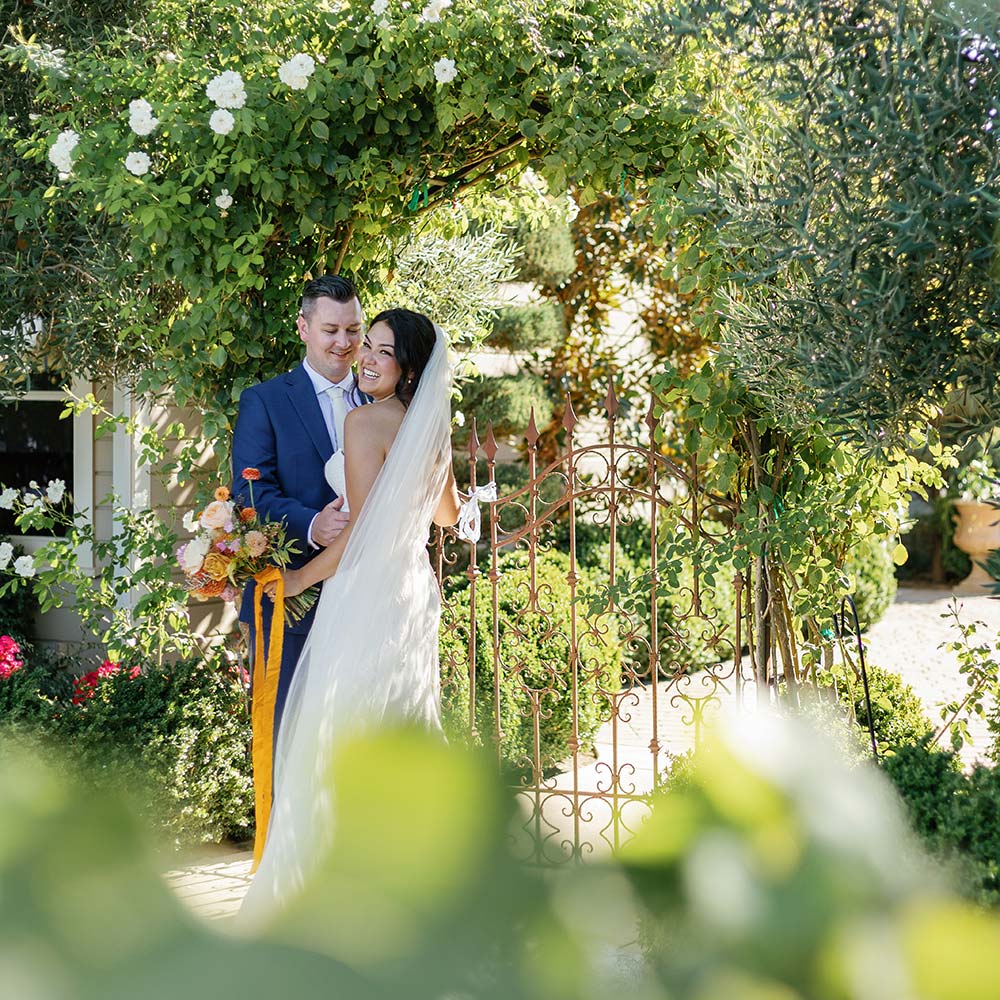 Peeking through the vines as the bride and groom pose in front of the winery and wrought iron fence at their outdoor winery wedding in Sacramento.