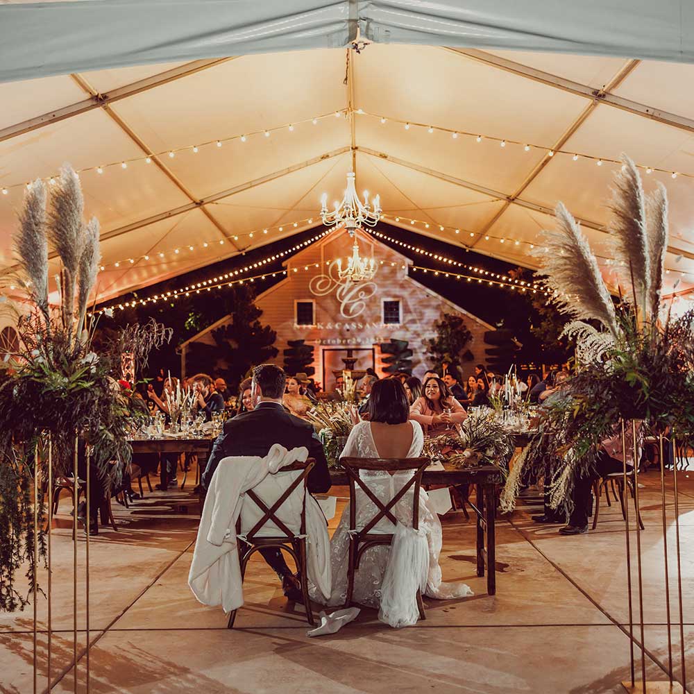 This evening winery wedding is lit up with market lights under the tented winery wedding as we see the back of the bride and groom and faces of all of the guests with the 1918 winery in full view.