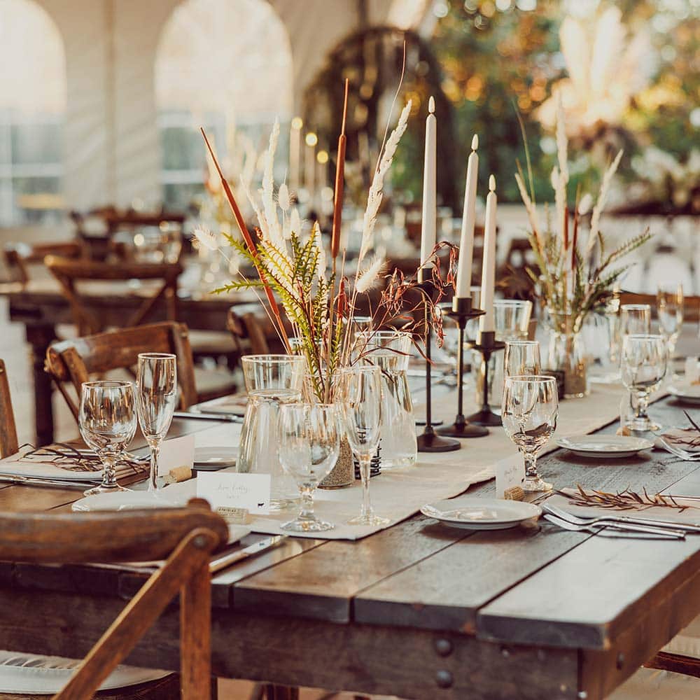 A decorated wedding guest table with simple white linens and candles and florals in the middle with the outdoor fireplace in the background under a covered tent at an premier outdoor winery wedding venue in the Sacramento area.