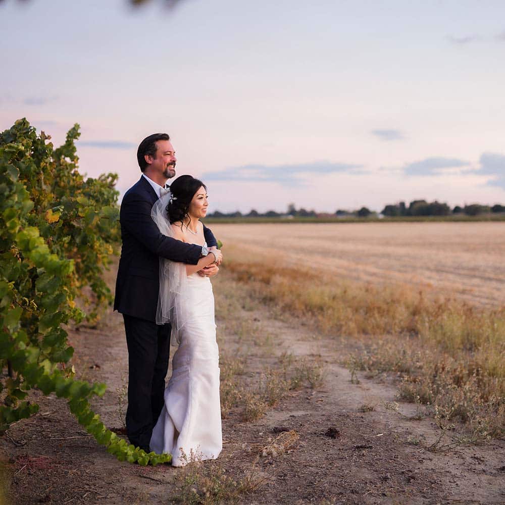 A groom embraces his bride in front of a vineyard looking out onto a harvested wheat field.