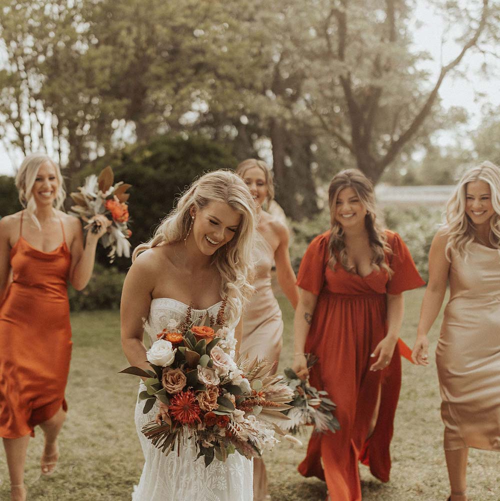 As they walk across the grass, a bride and her bridesmaids smile and laugh.