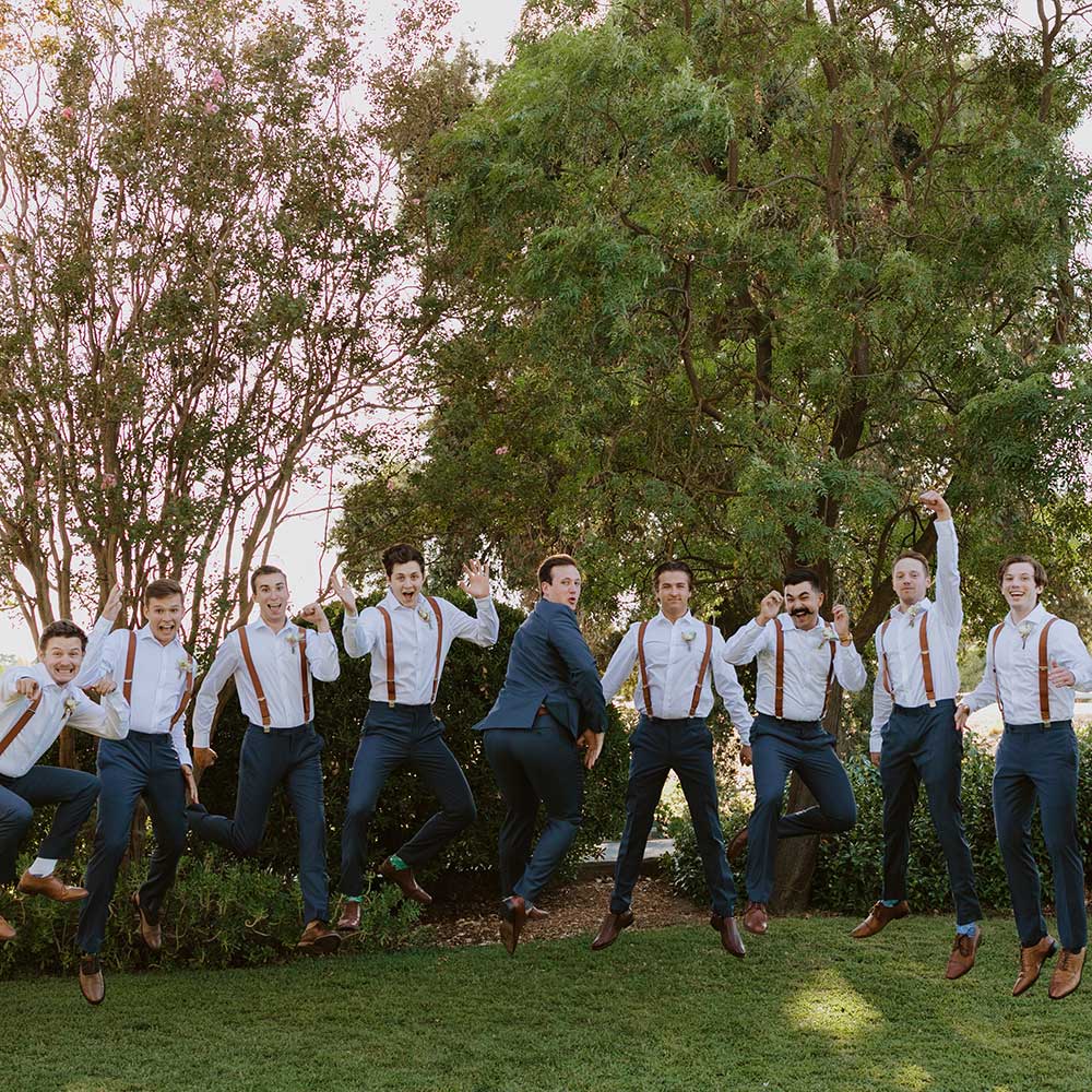 The groom and his groomsman jump in the air on the green grass with big smiles on their faces at their outdoor winery wedding in Sacramento.