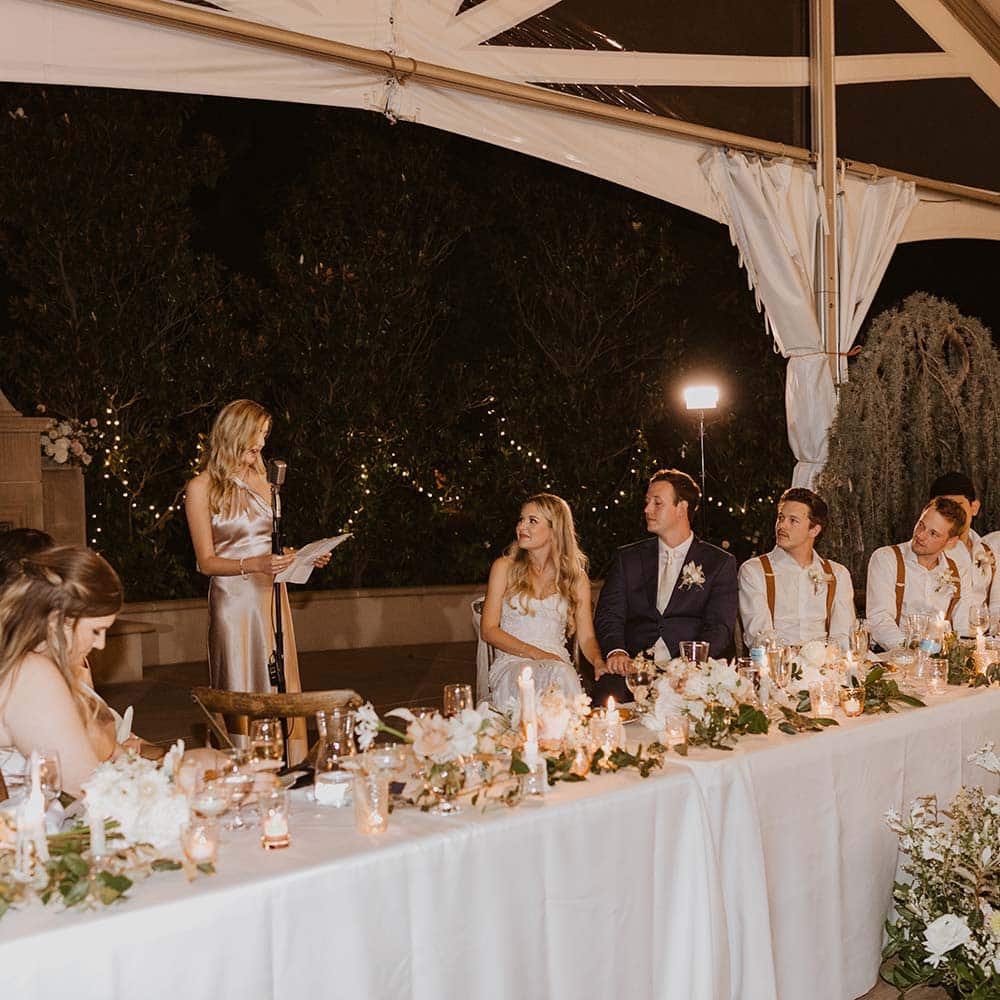 The bride and groom at their head table with their bridesmaids and groomsmen look onto the maid of honor giving her speech with a backdrop of market lights and an outdoor fireplace at this outdoor winery wedding in Sacramento.