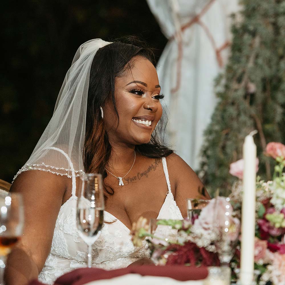 The smiling bride looks toward her guests while sitting at her head table which has floral centerpieces and candles at her outdoor winery wedding in Sacramento.