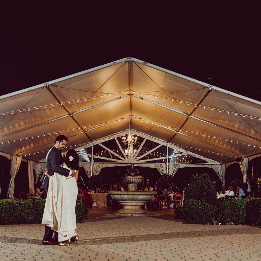 A night photo of the bride and groom having their first dance in front of the covered tent with market lights galore.