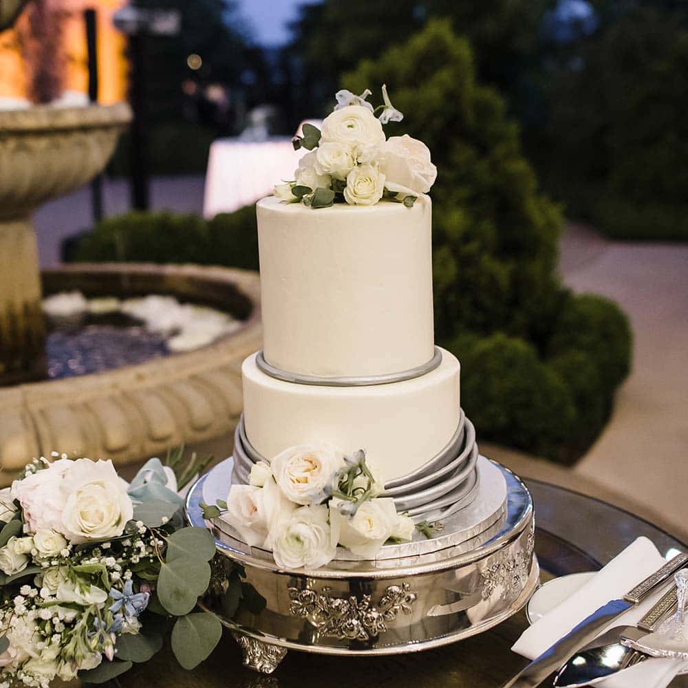 A two tiered cake sits on a glass top under a wine barrel in view of the three tiered fountain and market lights at this outdoor wedding venue.