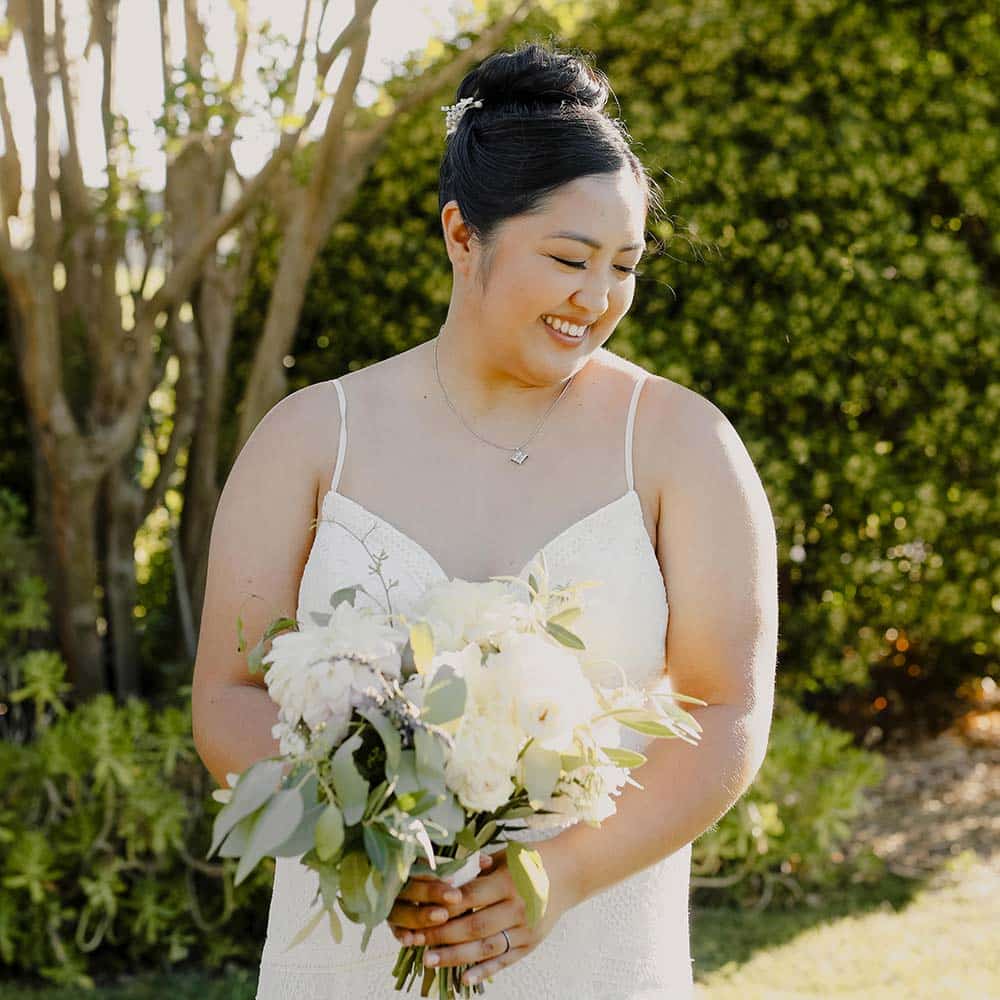 The brides smiles as she looks down and hold her white bouquet at her winery wedding in Sacramento.