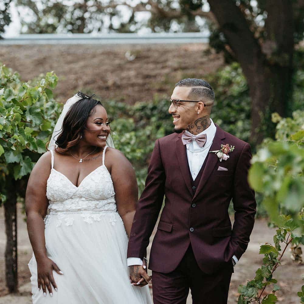 The bride in her white dress and veil and groom in his burgundy tuxedo hold hands smiling as they walk through the vineyard at their Sacramento winery wedding.