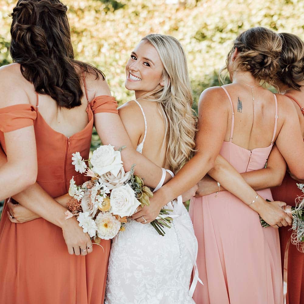 A bride and her bridesmaids have their back to the camera and their arms around each other as the bride only looks back at the camera.