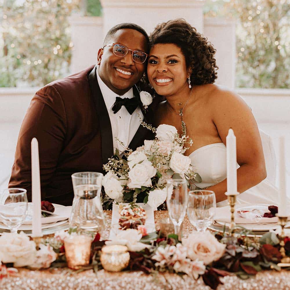 The bride and groom hold each other at their head table with is adorned with a gold glittery tablecloth, pink and burgundy flowers, candles and crystal glasses and they smile into the camera.