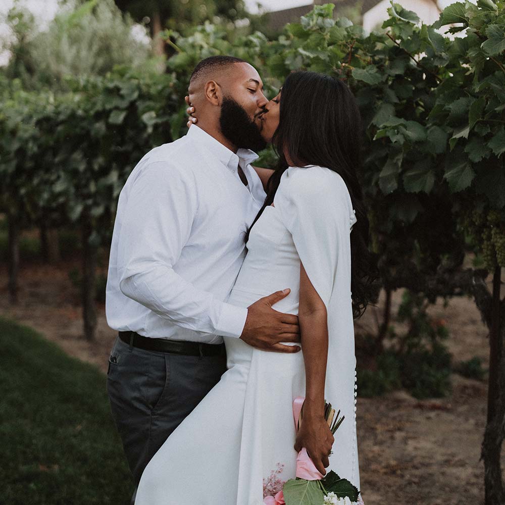 a groom holds onto his bride as they share a kiss as she hold onto her pink and white rose bouquet as the stand next to the chardonnay vines.