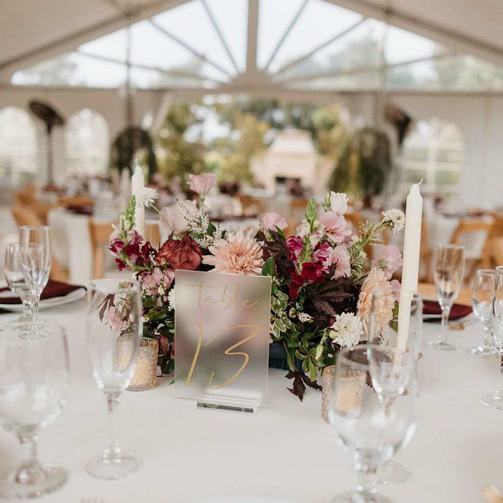 Table number 13 adorned with white linens and burgundy napkins with a pink and burgundy flower centerpiece and under a covered tent looking onto an outdoor fireplace at a winery in Sacramento.