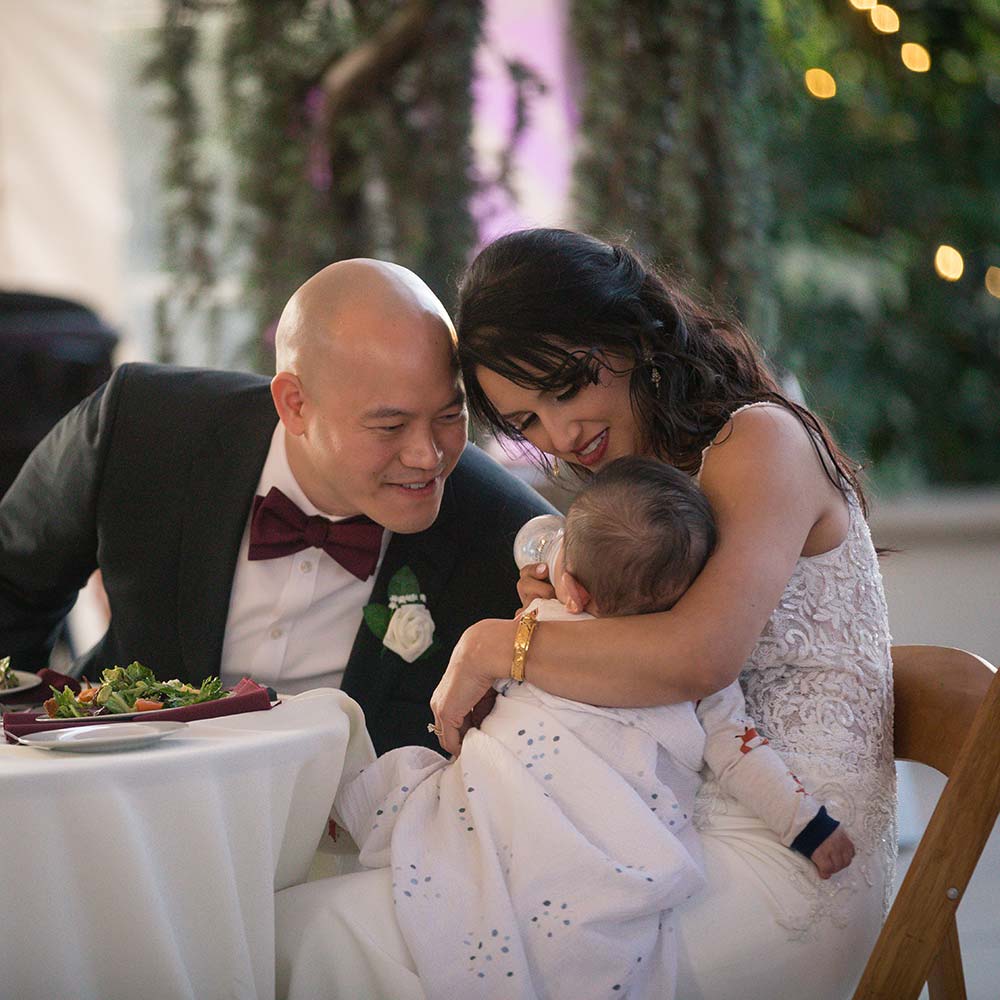The bride holds her baby at the head tables as the groom looks at the child with a loving smile.