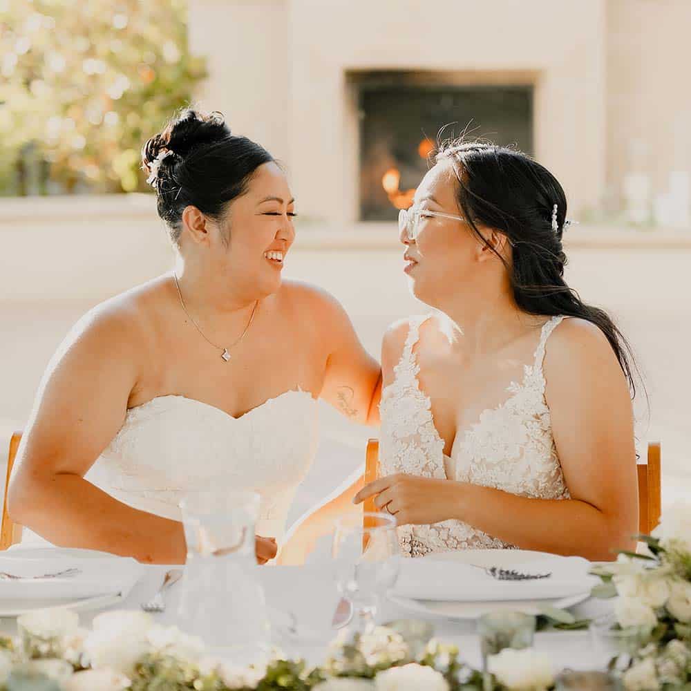 The two brides look at each other smiling as they sit as their head table with the lit outdoor fireplace in the background at their outdoor winery wedding.