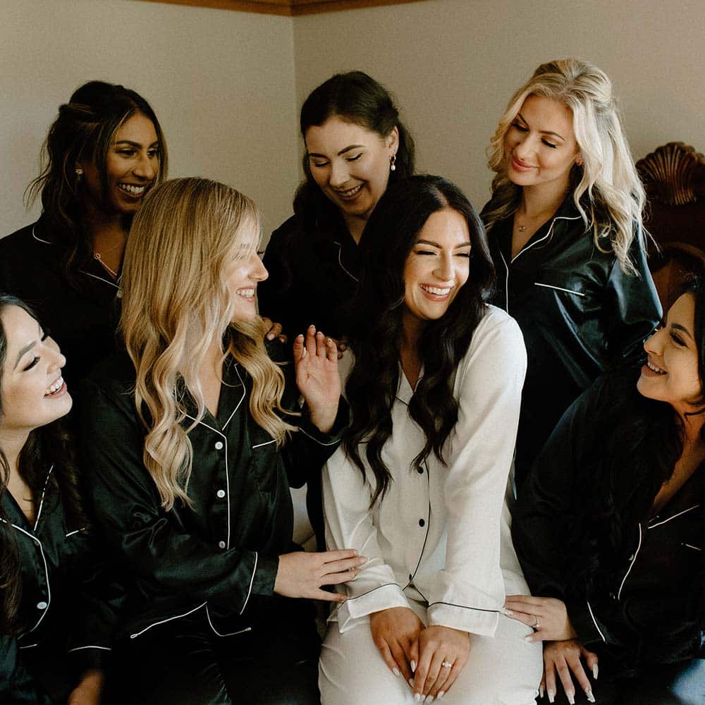The bride is in white silk pajamas and surrounded by her bridesmaids in black silk pajamas as they sit on a bed and all smile at the bride.