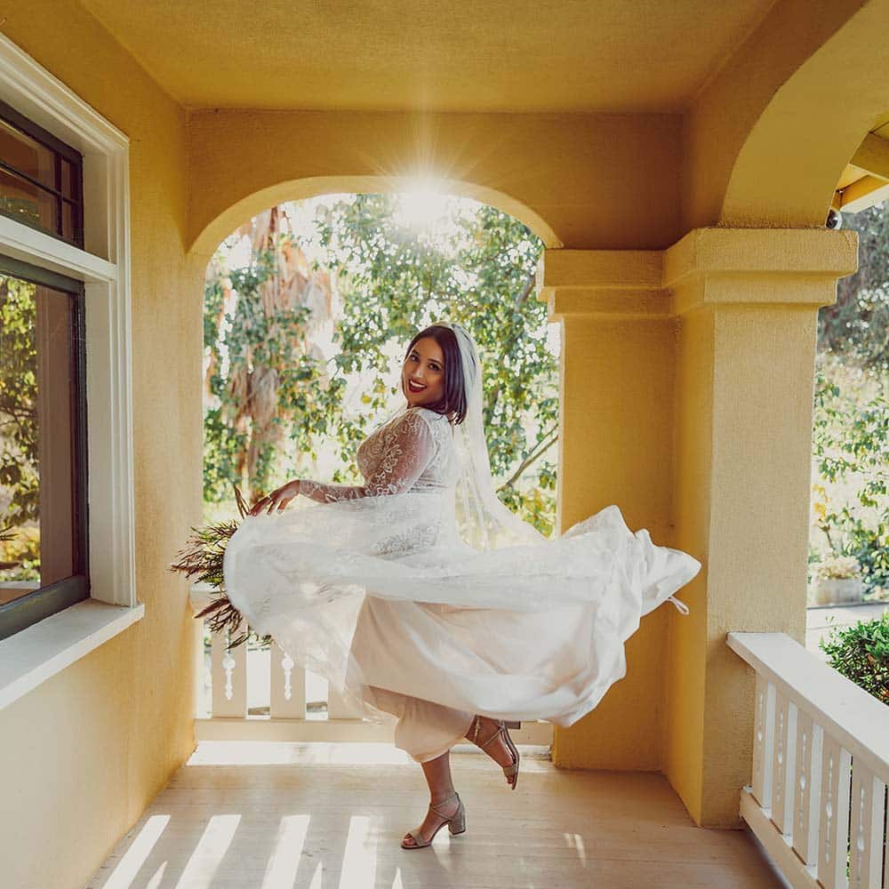 The brides twirls around in her white wedding dress as she holds her bouquet loosely in her hand on the outdoor porch of a 1920's craftsman home as the sun just starts to fall.