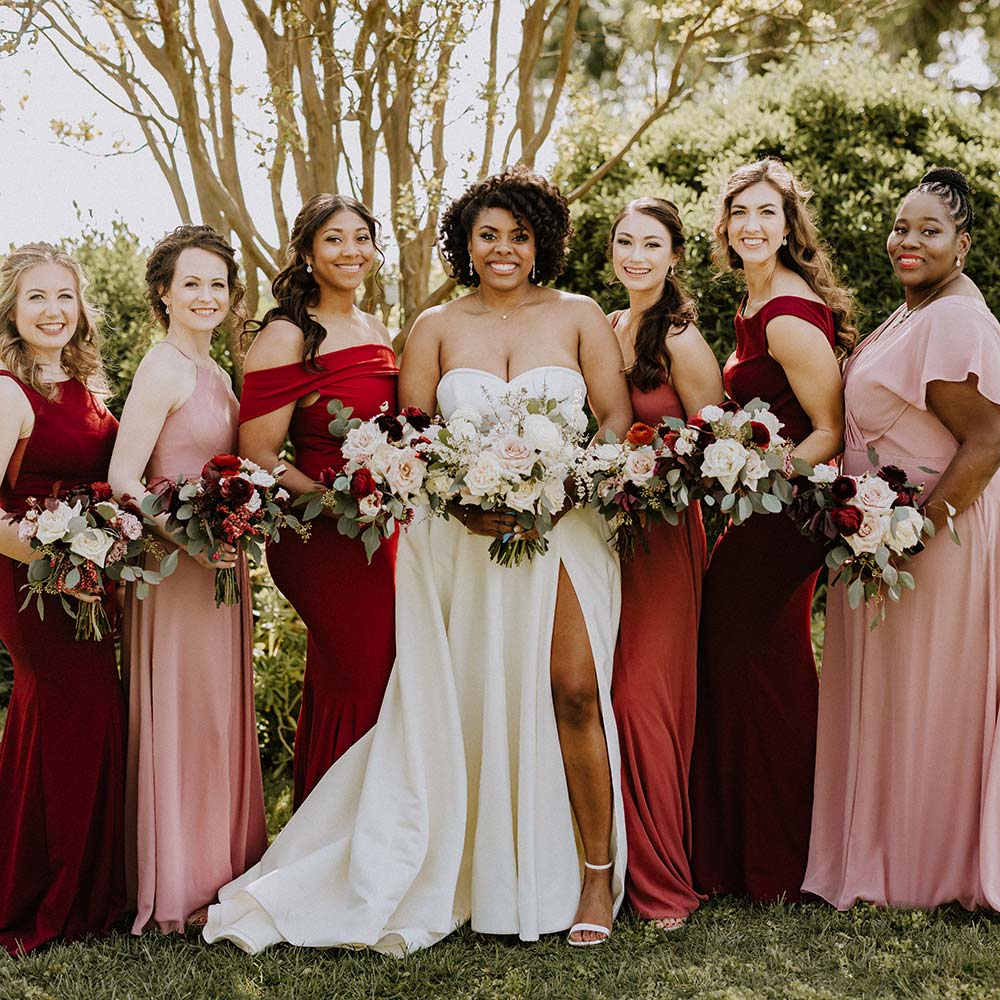 Surrounded by lush green mature trees, the bride and her bridal party smile into the camera at their outdoor winery wedding.