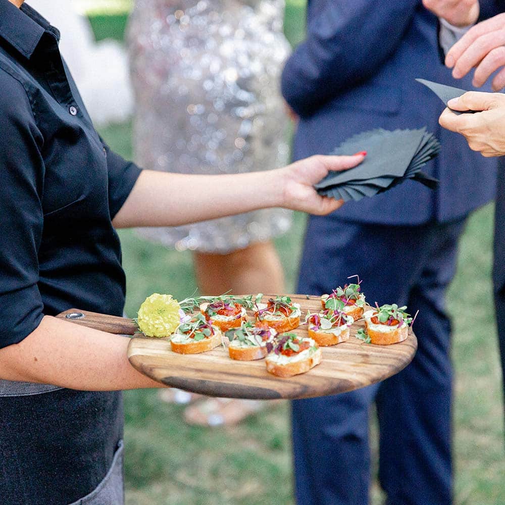 A server hold black napkins in one hand and a wooden board with appetizers in the other and offers them to the guests.