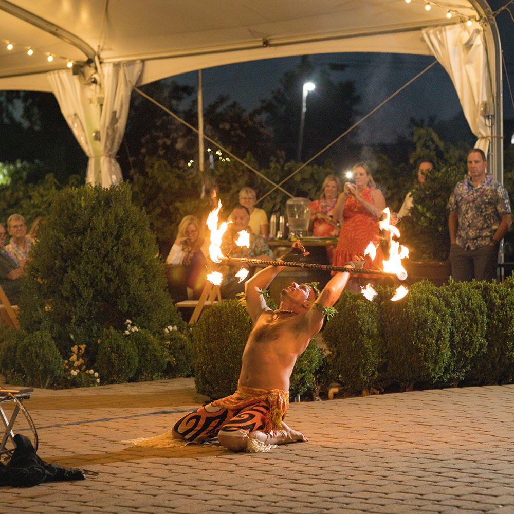 A fire dancer on kneeling knees hold his baton with fire on each end of it as the crowd looks toward him.