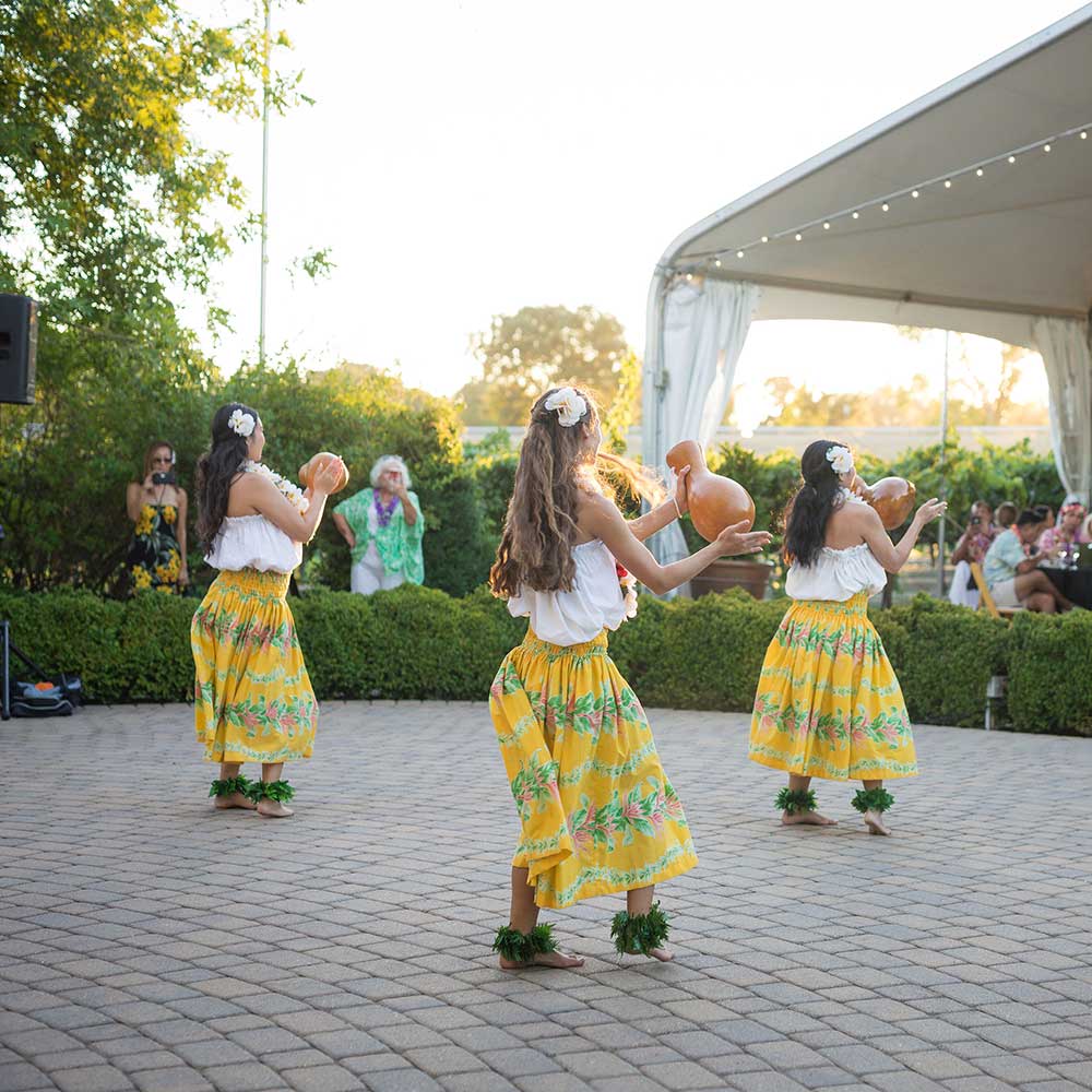 Three hula dancers performing for a group sitting under a tent.